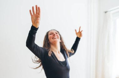 Siyah kıyafetli neşeli kadın dans ederken yakalandı. Parlak oda kollarında duygusal olarak yükseldi. Kıvırcık saçları özgürce akıyor ve yüzü mutluluk ve enerji saçıyor hareketin ve özgürlüğün özünü somutlaştırıyor.