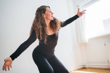 Siyah elbiseli bir kadın şık bir odada zarif bir şekilde dans ediyor. Hareketleri akıcı ve anlamlı. Kollarını uzatırken kıvırcık saçları uçuşuyor, dansında zarafet ve tutkuyu somutlaştırıyor.