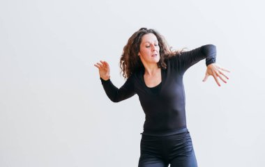 Siyah elbiseli kadın, omuzlarının üzerinde kıvırcık saçlarıyla dans ederken neşe içinde gülüyor. Etkileyici hareketler ve ışıl ışıl gülümseme. Saf mutluluğun görüntüsü. Minimalist ortamda özgürlük.