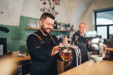 Siyah üniformalı şık sakallı barmen kameraya gülümsüyor, bar tezgahına bira vuruyor ve garsonu tepsiyle dürtüyor. Başarılı insanlar takım çalışması, arkadaşlık, demleme ve restoran endüstrisi konsepti imajı