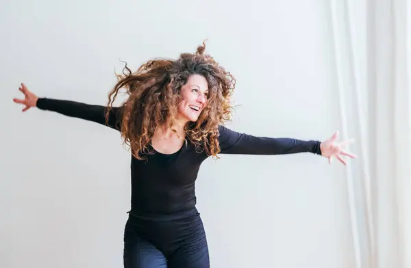stock image woman in black outfit laughs joyfully while dancing curly hair cascading over her shoulders. Expressive movements and radiant smile capture moment of pure happiness, freedom in minimalist setting
