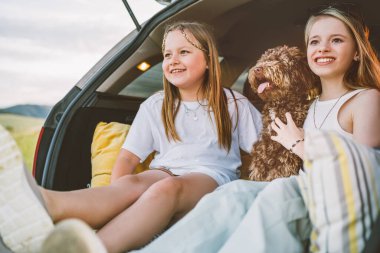 Küçük kızlar sen arabanın bagajında otururken pofuduk kahverengi Maltipoo köpeğiyle oynuyorlar. Otomobil yolculuğu sırasında neşeli ve canlı bir sahne yaratmak için köpekle oynuyorlar. Modern aile kavramında komik hayvanlar.
