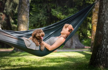 Güneşli bir günde hamakta tüylü kahverengi köpeğiyle uyuyan bir kadın. İkisi de mutlu ve mutlu görünüyor. Bu sakin açık hava sahnesi evcil hayvanlarla bağ kurmanın ve doğanın tadını çıkarmanın keyfini çıkarıyor..