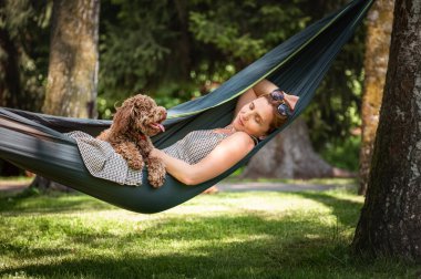 Kadınlar güneşli bir günde hamakta yumuşak kahverengi köpeğiyle rahatlar. İkisi de mutlu ve mutlu görünüyor. Bu sakin açık hava sahnesi evcil hayvanlarla bağ kurmanın ve doğanın tadını çıkarmanın keyfini çıkarıyor..