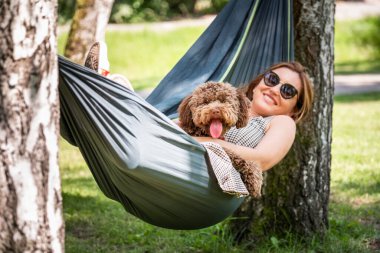 Kadınlar güneşli bir günde hamakta yumuşak kahverengi köpeğiyle rahatlar. İkisi de mutlu ve mutlu görünüyor. Bu sakin açık hava sahnesi evcil hayvanlarla bağ kurmanın ve doğanın tadını çıkarmanın keyfini çıkarıyor..