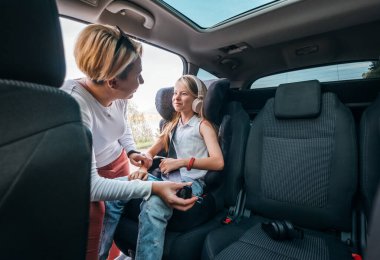 Inside the car photo of a mother fastening with safety auto belt her little daughter girl sitting in child seat. Girl listening music using headphones. Family values, traveling and technology concept clipart