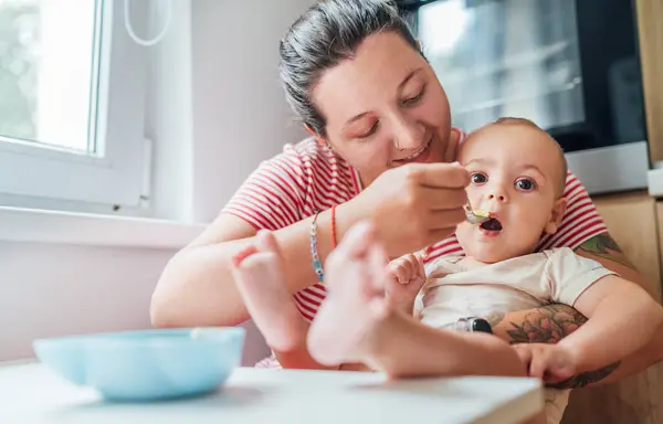 stock image Mother lovingly feeds her baby in cozy kitchen showcasing moment of tender care and bonding. Baby, with feet playfully extended, looks content and curious, adding touch of warmth and intimacy to scene