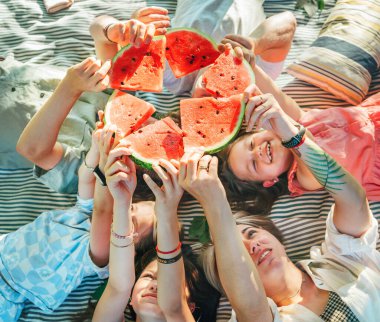 Hafta sonu güneşli bir günde aileleriyle piknik battaniyesine uzanmış gülümseyen, gülen ve sulu kırmızı karpuz parçaları yetiştiren genç kızlar. Aile değerleri, meyve vitaminleri, açık hava zamanı konsepti.