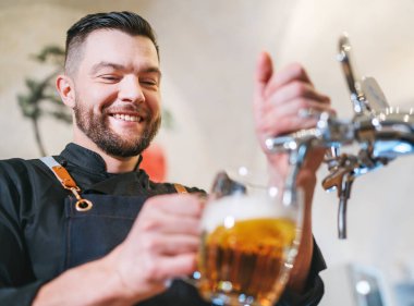 Gülümseyen şık sakallı barmen, siyah üniformalı, önlüklü barmen bardaki bardakta yeni bira içiyor. Başarılı insanlar, bira tüketimi, içecek endüstrisi imajı