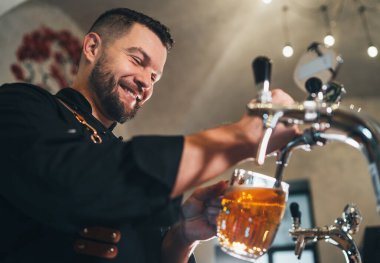 Gülümseyen şık sakallı barmen siyah üniformalı önlüklü barmen bardaki cam bardağa bira tıklatıyor. Başarılı insanlar, bira tüketimi, içecek endüstrisi imajı