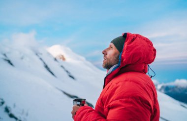 Portrait of high altitude mountaineer dressed red warm dawn jacket holding metal mug of hot tea thinking and enjoying fresh mountains frosty air. Mera Peak High Camp 5700m, Himalayas, Nepal. clipart