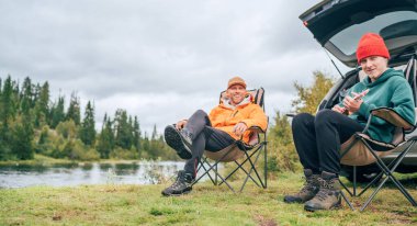Göl kenarında baba ve oğulla huzurlu bir Norveç kamp sahnesi. Oğulları doğayla iç içe oturmuş ukulele çalarken, onlar koltuklarda dinleniyorlar. Arabalarının yanında eğleniyorlar.