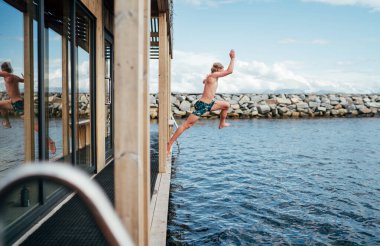 İskandinav yüzen sauna güvertesinden soğuk fiyort suyuna atlayan genç bir çocuk. Mutluluk ve rahatlık içinde parıldıyor. Huzurlu ve canlandırıcı sauna deneyimini mükemmel bir şekilde yakalıyor.