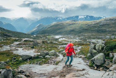 Kadın yürüyüşçü, Norveç dağlarında Trolltunga yürüyüş rotasının keyfini çıkarırken, parlak kırmızı ceketli ve yürüyüş direkleri olan bir yürüyüşçü..