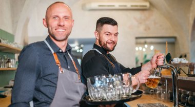 Tarz sahibi sakallı barmen siyah üniformalı barmen bar tezgahına tıklatıyor ve garsonla sohbet ediyor. Birbirlerine gülümsüyor.