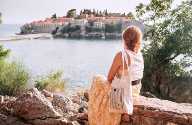 Karadağ 'da Sveti Stefan' ın keyfini çıkaran turist kadın, ikonik kırmızı çatılı binalarla dolu lüks bir otel adasını resmediyor. Adriyatik kıyısındaki lüks tatil beldelerinin ayrıcalığı ve seyahat konsepti imajı