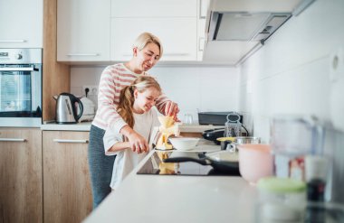 Anne ve kızı modern mutfakta birlikte sıcak vakit geçirip yemek hazırlıyorlar. Kızları kıyma makinesi kullanırken sıcak bir şekilde gülümsüyor. Aile bağlarını ve aile hayatını resmetmek için mükemmel. 