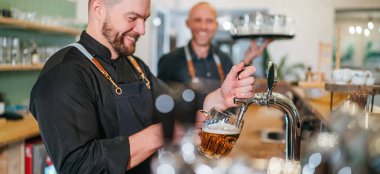 Tarz sahibi sakallı barmen siyah üniformalı barmen bar tezgahına tıklatıyor ve garsonla sohbet ediyor. Birbirlerine gülümsüyor.