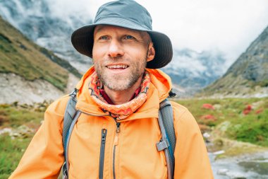 Portrait of smiling Man with backpack dressed orange waterproof jacket and funny hat walking the path during Makalu Barun National Park trek in Nepal. Mountain hiking and active people concept image. clipart