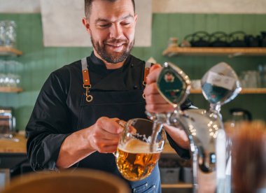 Gülümseyen şık sakallı barmen siyah üniformalı önlüklü barmen bardaki cam bardağa bira tıklatıyor. Başarılı insanlar, bira tüketimi, içecek endüstrisi imajı