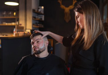 Uzun saçlı genç bayan berber, jilet gibi sakallı adamın yüzünü köpükle tıraş ettikten sonra kıllı kenarlara değer veriyor. Saç ve cilt bakımı, yerel küçük işletme konsepti..