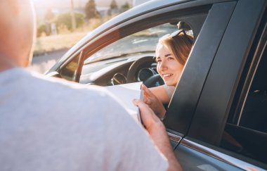 Young female sitting in car driver seat and sincerely smiling to deliveryman accepting two pizza boxes on the city street. Small business, people relations, fast foot meal consumption concept photo clipart