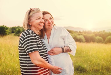 Portrait of a sincerely smiling young pregnant woman dressed in light summer clothes embracing with mother in evening sunset hours. Woman's health, human in the nature concept image. clipart