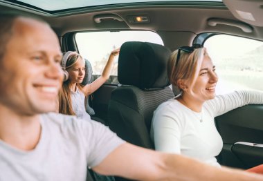 Happy young couple with daughter inside the modern car with panoramic roof during auto trop. They are smiling, laughing during road trip. Family values, traveling concepts.	
