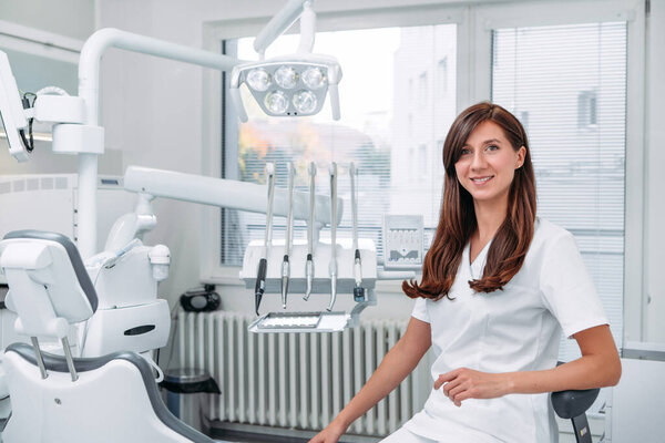 Portrait of sincerely smiling young dentist woman dressed white medical scrubs uniform sitting in modern dental clinic next to stomatology chair. Health care and medicare industry concept image.