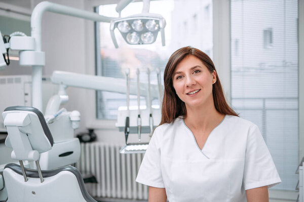 Portrait of sincerely smiling young dentist woman dressed white medical scrubs uniform sitting in modern dental clinic next to stomatology chair. Health care and medicare industry concept image.