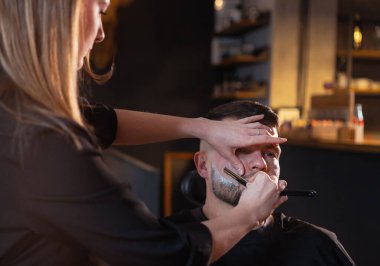 Uzun saçlı genç bayan berber sandalyedeyken sakallı adamın yüzünü nazikçe tıraş ediyor. Modern hafif siyah tarzı berber dükkanı iç mimarisi. Saç ve cilt bakımı, yerel küçük işletme konsepti..