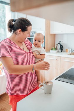 Anne, bebeğiyle sıcak bir mutfakta çay hazırlıyor. Nazik bir bakım ve bağ kurma anı. Kadın doğum izninde ve oğlunun birlikteliğinden zevk alıyor..