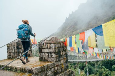 Askı Köprüsü 'nden geçen genç bayan yürüyüşçü, Lukla yakınlarındaki vadide asılı olan çok renkli Tibet Dua bayraklarıyla süslenmiş. Everest Merkez Kampı rotası, Sagarmatha Ulusal Parkı, Nepal
