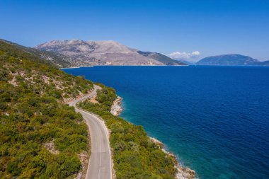 Sefalonya adasındaki Yunan kıyılarındaki sakin dalgaların yakınındaki deniz kıyısı kıvrımlı yolun insansız hava aracı görüntüsü. Ulaşım, doğal hava fotoğrafı.