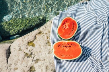 Kristal berrak su ve kayalık yüzeyin yanında çizgili kumaş üzerinde taze dilimlenmiş karpuz canlı yaz titreşimleri, doğal dokular ve açık havada canlandırıcı piknik atmosferi yaratır.