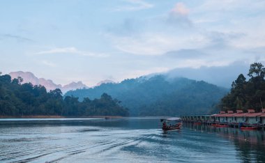 Yağmur ormanları ve akşam bulutlarıyla çevrili, inanılmaz Cheow Lan Gölü manzaralı yüzen köy kulübeleri. Surat Thani Eyaleti, Tayland 'da çarpıcı bir manzara..