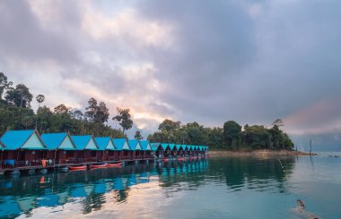 Floating village huts with incredible view of Cheow Lan Lake, surrounded by rainforest jungle and evening clouds. Stunning scenery in Surat Thani Province, Thailand. clipart