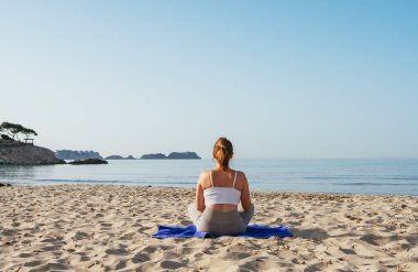 Sakin bir kadın sabahın erken saatlerinde sakin deniz dalgalarıyla kumlu sahilde oturup derin nefes egzersizleri yapıyor ve meditasyon yapıyor. Akıl sağlığı, dinlenen insanlar, seyahat konsepti.
