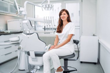 İçtenlikle gülümseyen genç dişçi kadın portresi beyaz önlük giymiş modern diş hekimliği kliniğinde stomatoloji kürsüsünün yanında oturuyor. Sağlık ve sağlık sektörü konsepti.