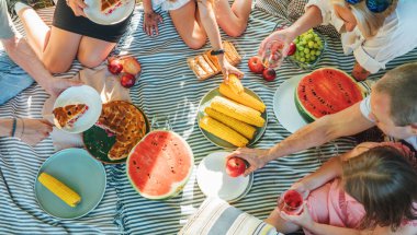 Şehir parkında piknik battaniyesinde oturan büyük aile manzarası. Haşlanmış mısır, elma, şeftali, hamur işi ve karpuz yiyorlar. Aile değerleri ve açık hava aktiviteleri kavramı
