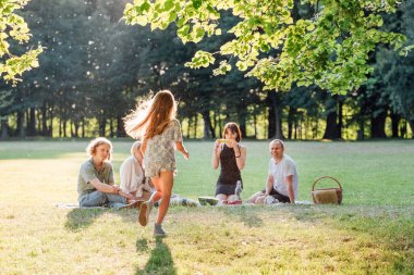 Yeşil şehir parkında ailesiyle piknik yaparken ailesine koşan yazlık elbiseli küçük uzun saçlı kız. Aile değerleri ve açık hava aktiviteleri kavramı