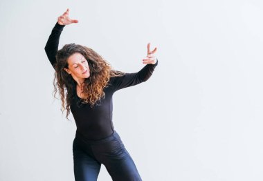 Siyah elbiseli kadın, omuzlarının üzerinde kıvırcık saçlarıyla dans ederken neşe içinde gülüyor. Etkileyici hareketler ve ışıl ışıl gülümseme. Saf mutluluğun görüntüsü. Minimalist ortamda özgürlük. .