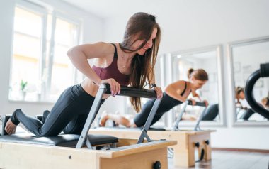 Spor spor salonundaki pilates reformatör spor aletini kullanarak uzun tahta statik kas güçlendirme egzersizi yapan iki genç bayan. Aktif insanlar eğitim, yoga dersleri konsepti