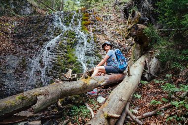 Sırt çantalı, şapkalı, aktif yürüyüş kıyafetleri ve botları olan gülümseyen bir kadın dağ nehri şelalesinin yanında oturuyor ve doğa gücünün tadını çıkarıyor. Seyahat, yürüyüş, doğa konsepti