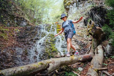 Aktif yürüyüş kıyafetleri giymiş sırt çantalı aktif bir kadın devrilmiş ağaç gövdesi köprüsünden geçen şelaleyle Mountain River Deresi 'ni geçiyor. Seyahat, yürüyüş, doğa konsepti