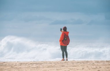 Portekiz 'in Porto kenti yakınlarındaki Aguda yerleşim yerindeki Praia da Aguda kumlu sahilinde, sırt çantalı yalnız bir kadın fotoğraf çekiyor ve kış fırtınasının tadını çıkarıyor.