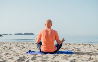 Sakin adam nilüfer pozisyonunda oturuyor, yoga yapıyor, derin nefes egzersizleri yapıyor ve sabahın erken saatlerinde sakin deniz dalgalarıyla kumlu sahilde meditasyon yapıyor. Akıl sağlığı, dinlenen insanlar, seyahat konsepti.