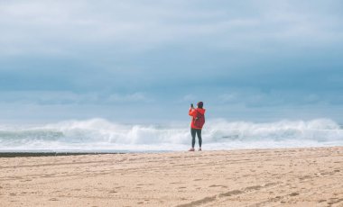 Portekiz 'in Porto kenti yakınlarındaki Aguda yerleşim yerindeki Praia da Aguda kumlu sahilinde, sırt çantalı yalnız bir kadın fotoğraf çekiyor ve kış fırtınasının tadını çıkarıyor.