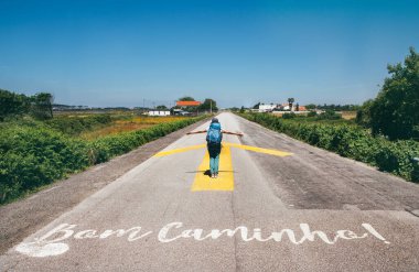 Sırt çantalı neşeli kadın hacı açık mavi gökyüzünün altında sarı ok yolunda yürüyor. Ünlü Camino de Santiago boyunca 