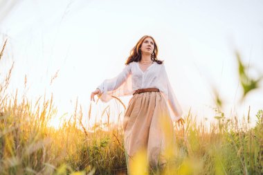 Beyaz bluz ve bej etekli genç bir kadın altın çayır boyunca özgürce yürüyor günbatımının keyfini çıkarıyor sıcak güneş ışığının barışçıl doğanın atmosferinin tadını çıkarıyor özgürlüğün farkındalığını ve doğayla uyum içinde olduğunu ifade ediyor.